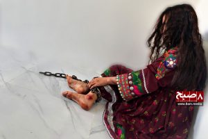 Une jeune Afghane est assise, enchaînée, ses pieds meurtris, témoignage silencieux du traumatisme du mariage forcé et des violences conjugales. Sa robe colorée contraste avec la sombre réalité de son enfermement. (Crédit photo : 8am.media)