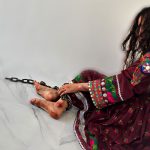 Une jeune femme afghane est assise enchaînée, ses pieds meurtris témoignant silencieusement du traumatisme du mariage forcé et de la violence domestique. Sa robe colorée contraste avec la triste réalité de son confinement. (Crédit photo : 8am.media)